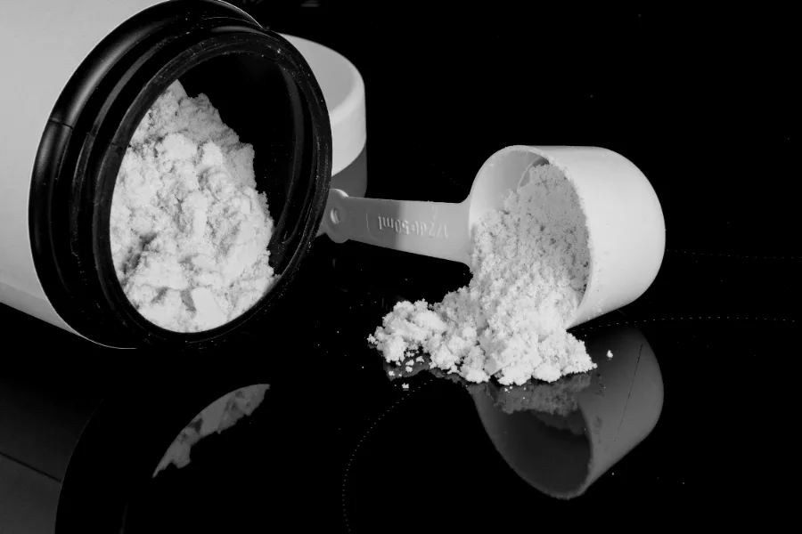 A tub of protein powder tipped over beside a full scoop on a dark reflective surface.