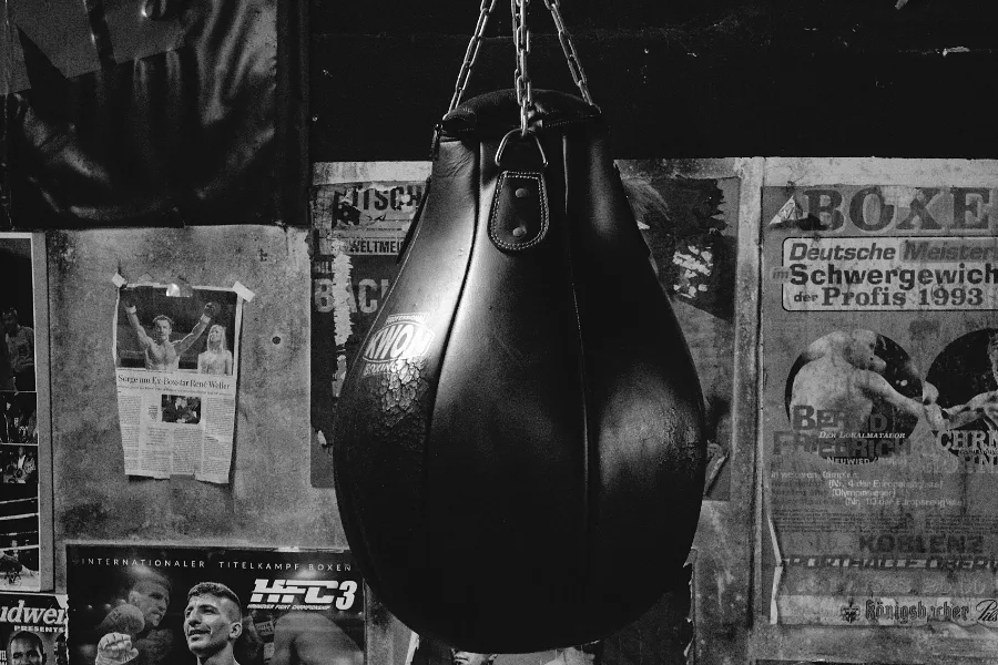 A worn leather heavy bag hanging in front of a wall of old fight posters.